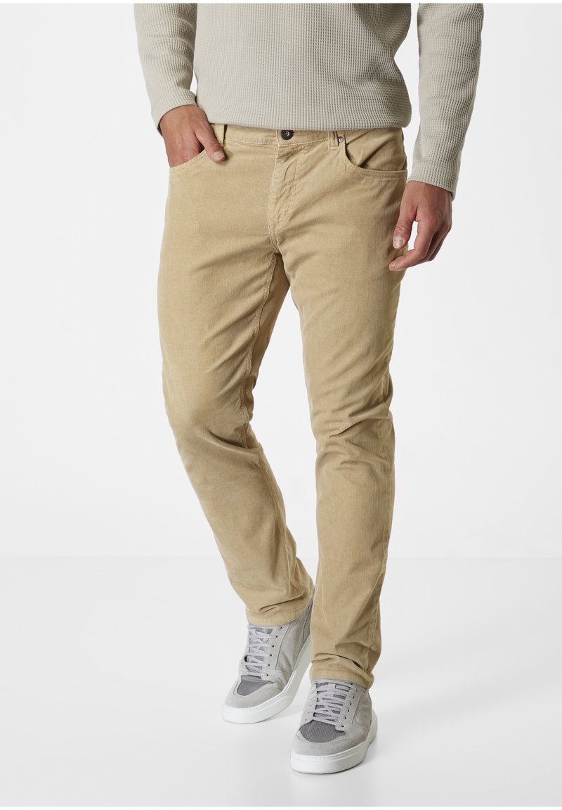 Beige Cordhosen mit schmaler Passform, ausgestattet mit Fronttaschen und einem Knopfverschluss. Kombiniert mit hellgrauen Sneakers für einen lässigen Look.