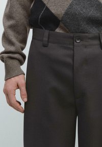 Dunkelgraue Hose aus glattem Stoff mit vorderer Knopfleiste und Seitentaschen, kombiniert mit einem beige gemusterten Pullover.