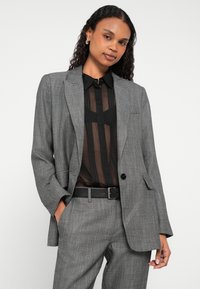 Szara marynarka w kratkę w jodełkę z podwiniętymi rękawami, w połączeniu z przejrzystą czarną bluzką i czarnym paskiem, zapinana na pojedynczy guzik.