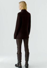 Donkerbruin gebreid sweater met een hoge hals, oversized ontwerp en textuurafwerking, gecombineerd met een nauwsluitende bruine leren broek en kniehoog laarzen.