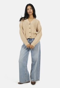 Cardigan beige texturé avec une fermeture à boutons, associé à un jean bleu clair à jambes larges. Des chaussures à imprimé léopard remarquables complètent la tenue.