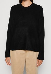 Personne portant un pull en maille noir à manches longues et un pantalon beige, debout devant un fond gris clair uni.