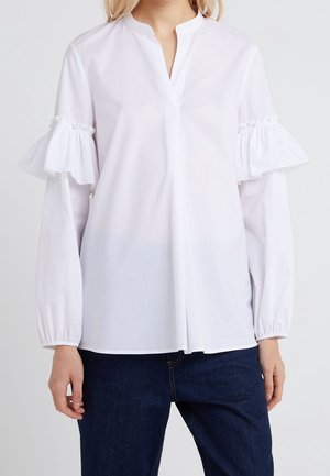 Kvinde iført en hvid bluse med flæseudskårne ærmer og elastiske manchetter, kombineret med mørkeblå denimjeans, stående mod en ensfarvet baggrund.