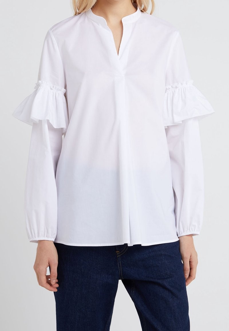 Femme portant un chemisier blanc à manches découpées volantées avec poignets élastiques, associé à un jean en denim bleu foncé, debout devant un fond uni.