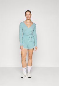 Svares tynn top i lys teal med lange ermer, kombinert med matchende shorts med elastisk liv. Generell avslappet passform, bomullslignende tekstur. Hvite sportssokker og sko.