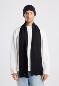 Conjunto de gorro y bufanda de punto acanalado negro, hecho de material suave. La bufanda tiene forma rectangular larga con una etiqueta cerca del extremo.