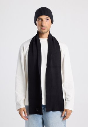 Zwart geribbeld gebreid beanie en sjaalset, gemaakt van zachte materialen. De sjaal heeft een lange rechthoekige vorm met een label nabij het einde.
