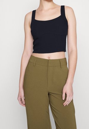 Woman wearing a black cropped tank top with wide straps and high-waisted olive green pants against a plain background.