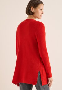 Roter gestrickter Pullover mit langen Ärmeln, seitlichen Schlitzen und einem lockeren Schnitt, aus einem strukturierten Stoff gefertigt. Keine weiteren sichtbaren Details.