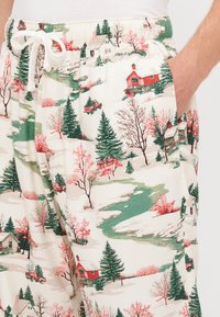 Pamut pizsama nadrág téli tájat ábrázoló mintával, vörös házakkal, zöld fákkal és rózsaszín virágokkal, krémszínű háttér előtt.