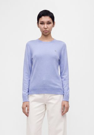 Femme aux cheveux courts et foncés portant un pull violet clair à manches longues et un pantalon blanc, debout devant un fond clair uni.