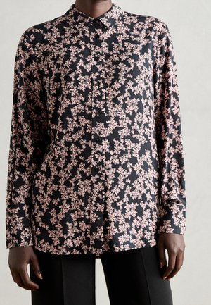 Persoon draagt een zwart overhemd met lange mouwen en knopen, voorzien van een roze abstract bloemmotief, en een zwarte broek, staand tegen een effen achtergrond.