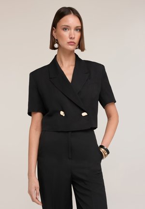 Kvinde med glat brunt hår bærer sort cropped dobbeltbreasted blazer med guldknapper, matchende højtaljede bukser, perleøreringe og armbånd.