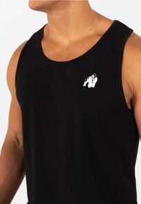 Gespiereerd persoon draagt een zwarte mouwloze sportshirt met een klein wit gorillalogo op de linkerbovenkant van de borst tegen een effen achtergrond.