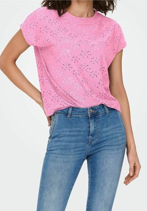 Femme portant un haut en dentelle œillet rose à manches courtes, rentré dans un jean bleu taille haute, debout avec une main sur la hanche.