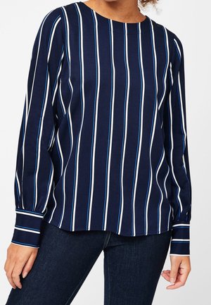 Kvinde iført en navyblå bluse med lodrette blå og hvide striber og lange ærmer, kombineret med mørkeblå jeans.
