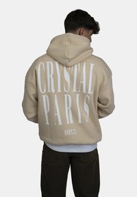 Felpa beige con una grande stampa bianca che recita "CRYSTAL PARIS" sul retro, caratterizzata da una vestibilità comoda e un cappuccio regolabile.
