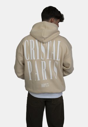 Beige hoodie met een grote witte opdruk met de tekst "CRYSTAL PARIS" op de rug, met een relaxed fit en een capuchon met trekkoord.