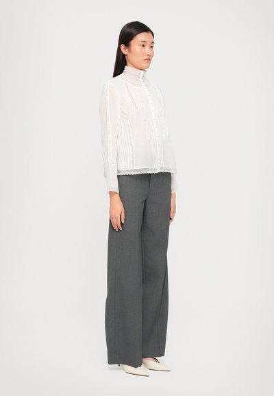 Blusa blanca de encaje con cuello alto y botones en el frente, presenta rayas verticales y patrones intrincados. Combinada con pantalones grises de pierna ancha.