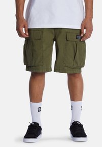 Bermudas cargo de color verde oliva con múltiples bolsillos laterales, con una tela texturizada y un ajuste relajado, llevadas con zapatos negros y calcetines blancos.