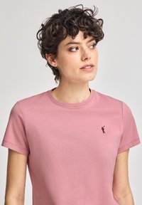 Camiseta de manga corta rosa en tela suave con un pequeño detalle de logotipo oscuro en el lado izquierdo del pecho. Cuello redondo y corte relajado.