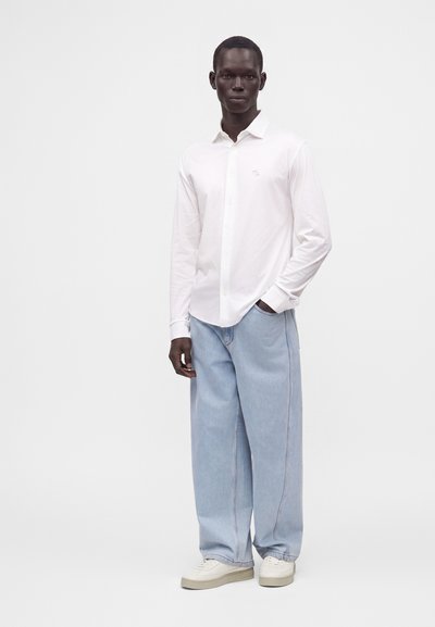 Homme portant une chemise blanche à manches longues boutonnée, un jean large bleu clair et des baskets blanches, debout devant un fond uni.