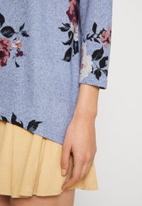 Ljusblå stickad tröja med trekvartslånga ärmar som har blommönster i burgunder och vitt, kombinerad med en veckad gul kjol.