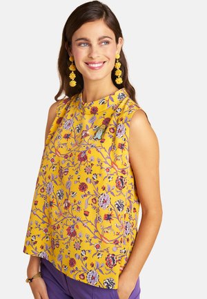 Smiling woman with long brown hair wears yellow floral sleeveless top, matching yellow floral earrings, and purple pants.