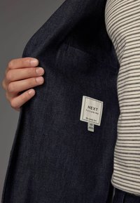 Dunkelblauer Denimjackeninnenraum mit einem Etikett, das "NEXT TAILORING RELAXED FIT" lautet. Sichtbare Taschen Details und ein gestreiftes Hemd darunter.