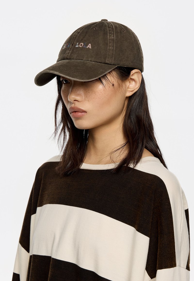 Gorra de color verde oliva oscuro con visera curva y logo bordado "ANAN LOLA" en colores contrastantes. Combinada con una camiseta a rayas en tonos marrón y crema.