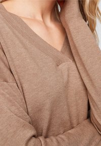 Sweter w serek z miękkiej, jasnobrązowej dzianiny. Posiada luźny krój z prążkowanymi mankietami i dekoltem, prezentując gładką, teksturowaną tkaninę.