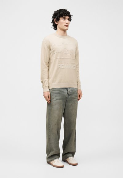 Νεαρός με κατσαρά μαλλιά φοράει μπεζ φούτερ Peanuts, φαρδιά ξεθωριασμένα πράσινα τζιν και λευκά αθλητικά παπούτσια, στέκεται μπροστά σε μια απλή φόντο.