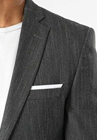 Blazer gris à motif avec un tissu texturé, présentant un revers cranté, un seul poche carrée blanche et un t-shirt blanc en dessous.