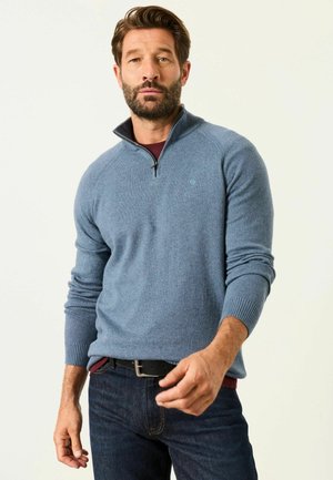 Mann mit Bart, der einen blauen Pullover mit Viertelreißverschluss und dunkle Jeans trägt, steht vor einem schlichten hellen Hintergrund und streckt die rechte Hand nach vorne.
