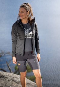 Vadderad svart jacka med ett grått innerfoder, kortärmad grafisk t-shirt och mörka shorts med dragkedjor. Bakgrund av vatten och träd.