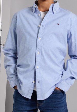 Man draagt een lichtblauw overhemd over een wit T-shirt, met handen in de zakken, staand tegen een effen achtergrond.