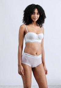 Νεαρή γυναίκα με σγουρά μαλλιά, φορώντας λευκό σουτιέν Calvin Klein και ανοιχτό ροζ εσώρουχα Calvin Klein, στέκεται μπροστά σε απλό ανοιχτό φόντο.