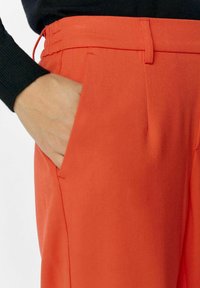Pantalons oranges avec une texture de tissu lisse, comportant des poches latérales et une taille ajustée. Un pull noir est porté sur le dessus.