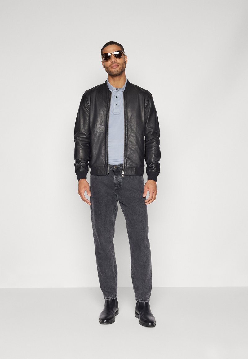 Veste bomber en cuir noir, polo bleu clair, jeans gris et bottes en cuir noir. Modèle portant des lunettes de soleil sombres, debout contre un arrière-plan neutre.