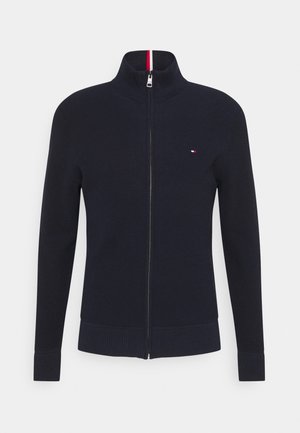 Pull en maille bleu marine à fermeture éclair intégrale, col haut, poignets et ourlet côtelés, avec un petit logo rouge, blanc et bleu sur la poitrine gauche.