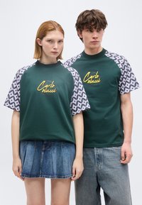 Δύο μοντέλα φορούν ταιριαστά σκούρα πράσινα t-shirts με ριγωτά μανίκια σε μωβ χρώμα και κίτρινο λογότυπο "Carlo Colucci", συνδυασμένα με τζιν παντελόνια.