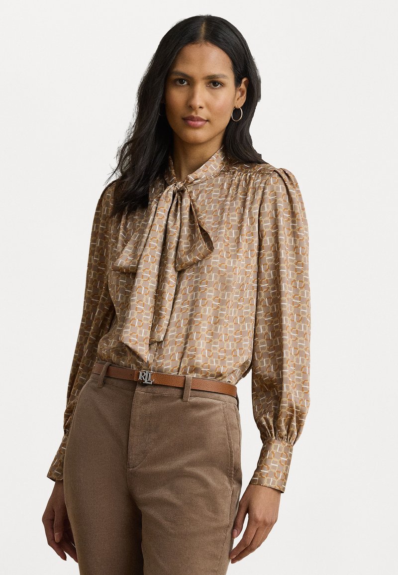 Blusa marrone con motivo a fantasia, colletto a fiocco, maniche ampie e un design geometrico sottile, abbinata a pantaloni in corduroy beige e una cintura marrone.