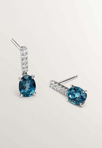 Los pendientes de plata presentan una gran gema azul ovalada en un engaste de garras, complementada por una fila de pequeñas piedras transparentes a lo largo del vástago.