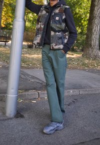 Chaleco de forro polar con estampado de camuflaje sobre una sudadera oscura, combinado con pantalones verdes y zapatillas grises. De pie junto a un poste en un entorno al aire libre.