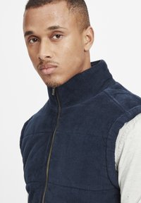 Jūras spēku krāsas korduroja veste ar augstu apkakli un rāvējslēdzēju. Tekstūrēta auduma un quilted dizains, uzlikta pāri gaiši pelēkai garajai piedurknei krekliņam.