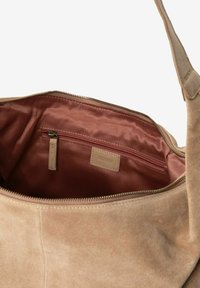 Intérieur d'un sac à bandoulière en suède beige avec une texture douce, comportant une doublure rose, une poche zippée et une plaque d'étiquette brodée.