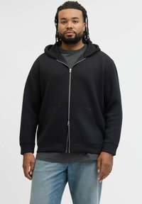 Felpa nera con zip, tasca frontale e cappuccio con coulisse, indossata sopra una maglietta grigia; il modello indossa jeans azzurri chiari.