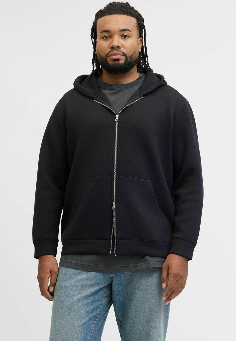 Felpa nera con zip, tasca frontale e cappuccio con coulisse, indossata sopra una maglietta grigia; il modello indossa jeans azzurri chiari.