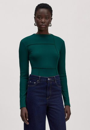 Vrouw met kort krullend haar, zilveren oorringen, een aansluitende groene top met lange mouwen en donkerblauwe hooggetailleerde jeans.