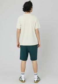 T-shirt crème à manches courtes associé à un short vert avec une poche arrière. Modèle portant des chaussettes blanches avec des rayures vertes et des baskets vertes.